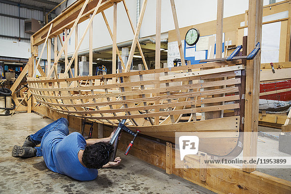 Auf dem Boden liegender Mann in der Werkstatt eines Bootsbauers  der an einem hölzernen Bootsrumpf arbeitet.
