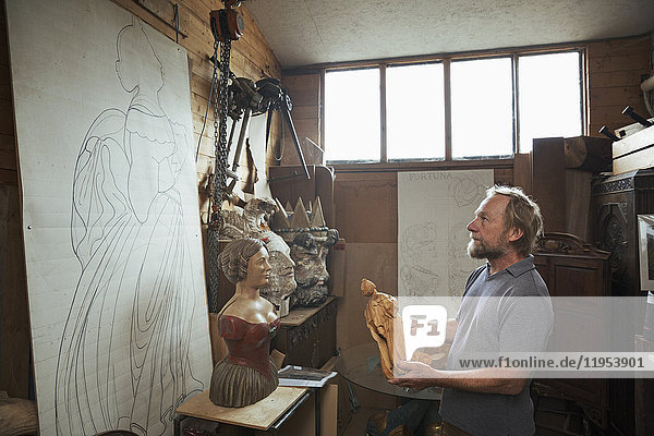 Ein handwerklicher Holzschnitzer hält eine kleine Holzfigur und betrachtet ein großes Muster an der Wand seiner Werkstatt. Geschnitzte hölzerne Schiffsfiguren.