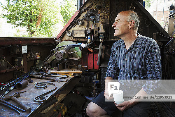 Blacksmith sitting on a working narrowboat on a waterway  in his workshop holding mug.