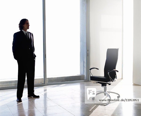 Businessman wearing dark suit standing indoors next to office chair in front of window  hands in pockets.