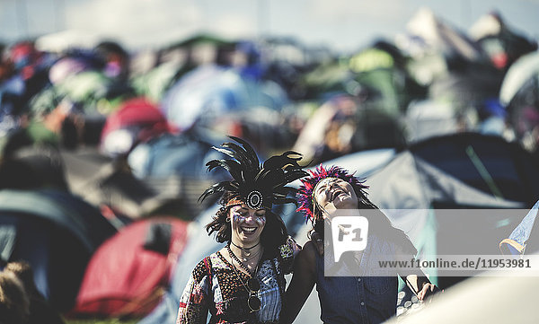 Two smiling young women at a summer music festival face painted  wearing feather headdress  standing among tents.