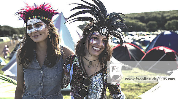 Zwei lächelnde junge Frauen bei einem Sommer-Musikfestival mit bemaltem Gesicht und Federkopfschmuck zwischen Zelten stehend.