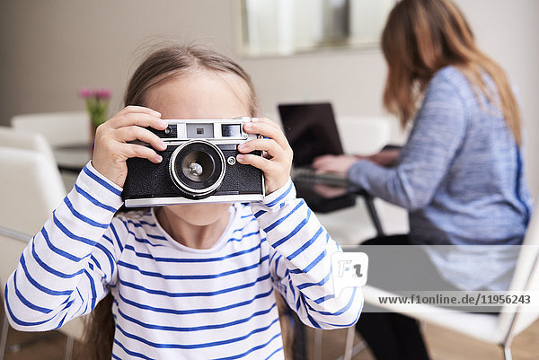 Little girl taking picture with camera while her mother working on laptop in the background