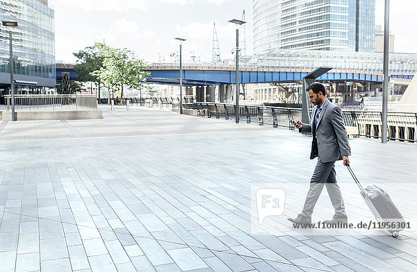 Businessman walking through the city with rolling suitcase and cell phone
