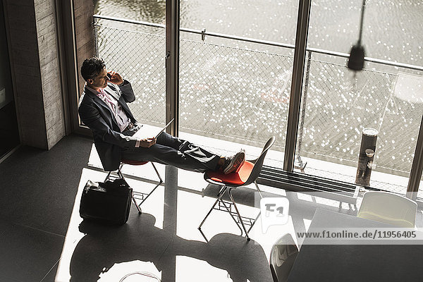 Geschäftsmann am Fenster sitzend mit erhobenen Füßen  am Laptop arbeitend und telefonierend