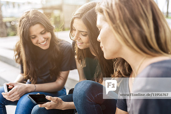 Drei glückliche junge Frauen sitzen draußen und schauen auf ihr Handy.