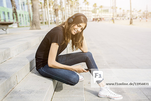 Lächelnde junge Frau sitzt auf einer Treppe und hört Musik.