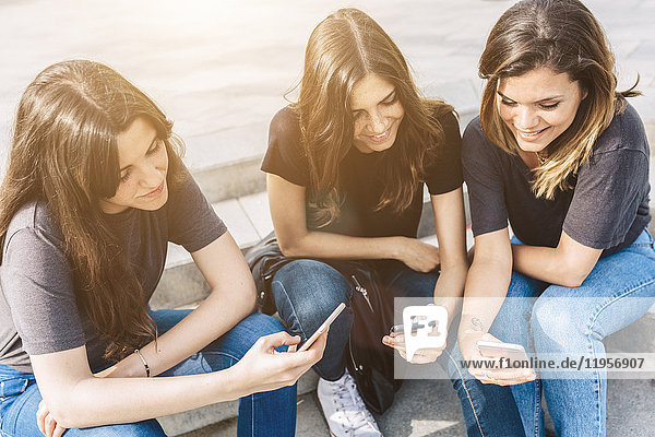 Drei lächelnde junge Frauen sitzen draußen und schauen auf Handys.
