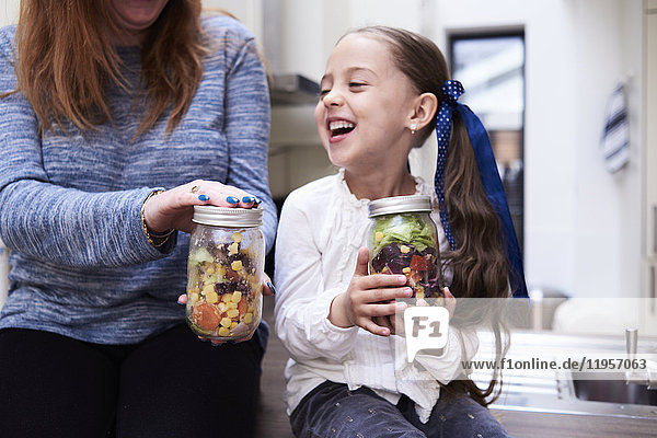 Lachendes kleines Mädchen mit einem Einmachglas mit gemischtem Salat neben ihrer Mutter.