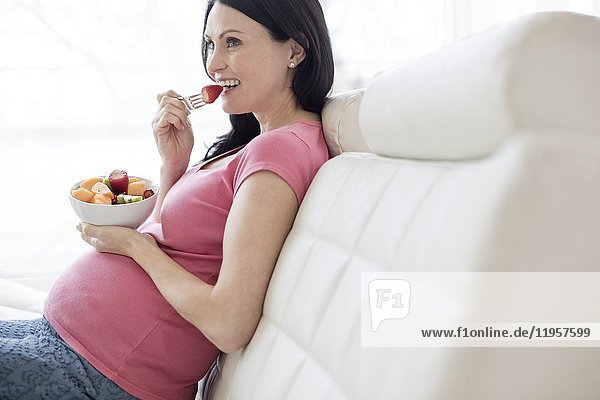 MODEL RELEASED. Pregnant woman on sofa eating fresh fruit.