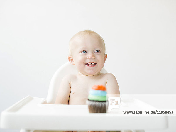Baby boy (12-17 months) sitting in high chair with cupcake in front of him