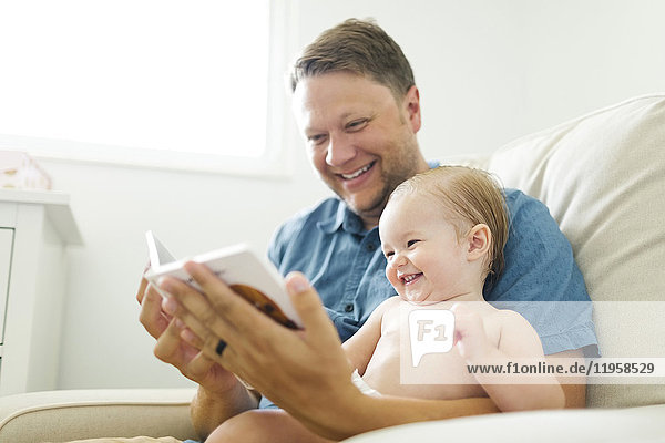 Vater mit einem kleinen Mädchen (12-17 Monate),  das im Wohnzimmer sitzt und ein Buch liest
