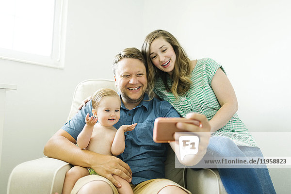 Mutter und Vater mit einem kleinen Mädchen (12-17 Monate) sitzen im Wohnzimmer und machen ein Selfie