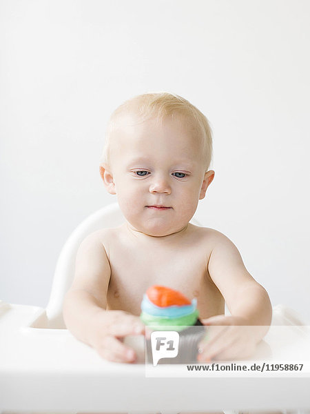 Baby boy (12-17 months) sitting in high chair and holding cupcake