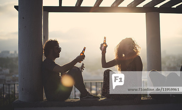 Silhouette eines Mannes und einer Frau  die bei Sonnenuntergang im Freien unter einer Pergola sitzen und Bierflaschen halten.