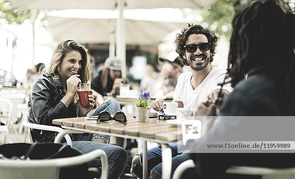 Zwei Männer und eine Frau sitzen draußen an einem Tisch in einem Café  lächeln und trinken.