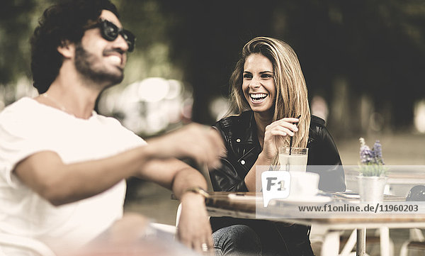 Bearded man wearing sunglasses and woman with long blond hair sitting outdoors at a table in a cafe  laughing.