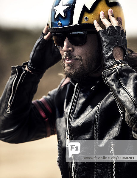 Bearded man wearing black leather jacket and sunglasses adjusting his yellow open face crash helmet.