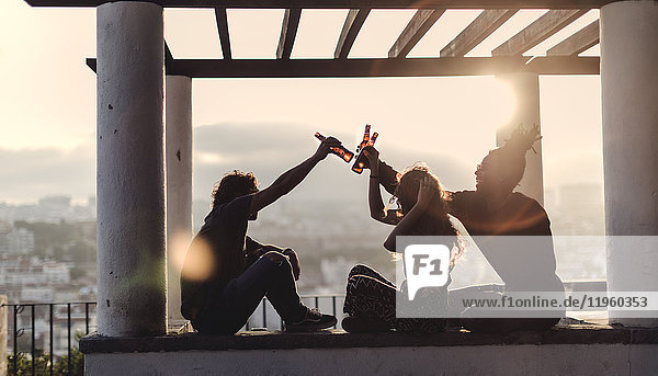 Silhouette von zwei Männern und einer Frau  die bei Sonnenuntergang im Freien unter einer Pergola sitzen  Bierflaschen in der Luft halten und auf den Sonnenuntergang anstoßen.