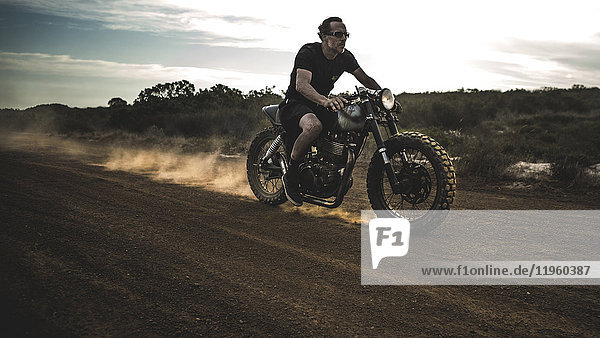 Man wearing shorts and sunglasses riding cafe racer motorcycle on a dusty dirt road.