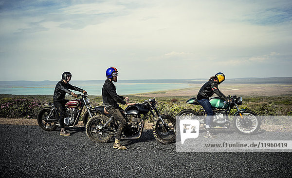 Three men wearing open face crash helmets sitting on cafe racer motorcycles on a rural road.