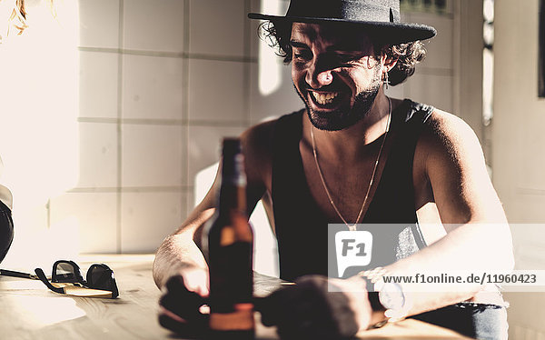 Bärtiger Mann mit Hut  der drinnen an einem Tisch sitzt  Bierflasche im Vordergrund.
