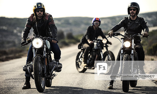Drei Männer mit offenen Sturzhelmen und Schutzbrille sitzen auf Cafe-Rennmotorrädern auf einer Landstraße.