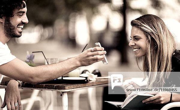 Bärtiger Mann mit Sonnenbrille und Frau mit langen blonden Haaren sitzt draußen an einem Tisch in einem Cafe  Frau schaut auf Smartphone  das der Mann in der Hand hält.