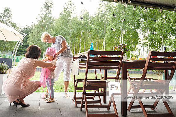 Senior and mature women dressing female toddler in pink bonnet at family lunch on patio