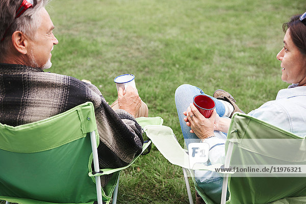 Erwachsenes Paar sitzt auf Campingstühlen  hält Blechbecher  Rückansicht