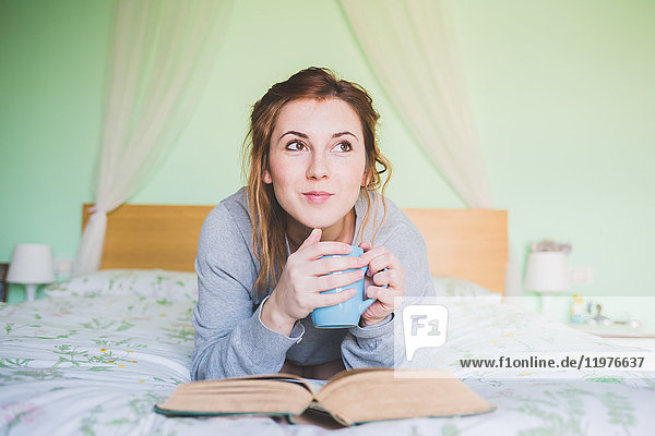 Junge Frau liegt auf dem Bett  hält Kaffeetasse und schaut auf