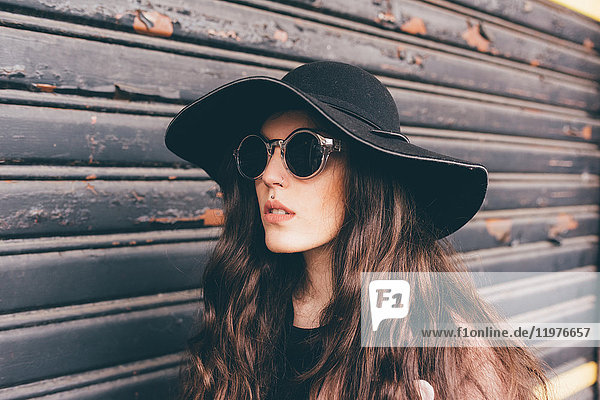 Portrait of young woman standing beside shutter  wearing floppy hat and sunglasses
