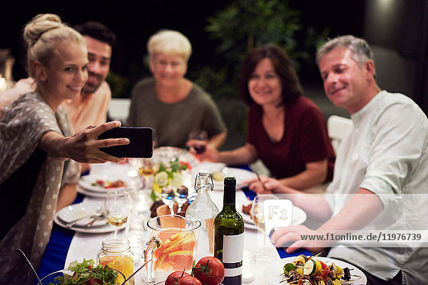 Group of people sitting at table  enjoying meal  young woman taking selfie of group using smartphone