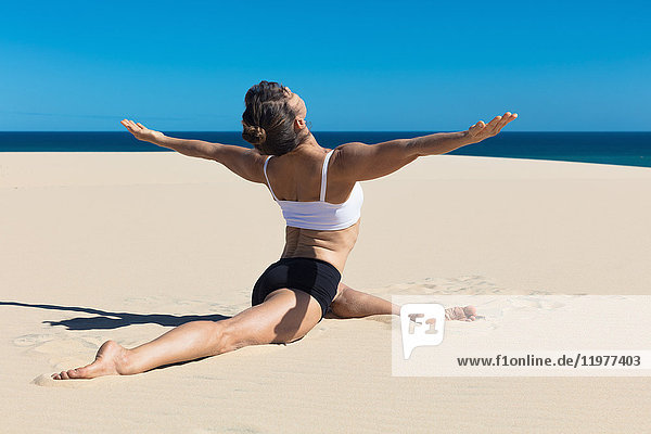 Rückansicht einer Frau am Strand beim Spagat,  Arme in Yogastellung geöffnet