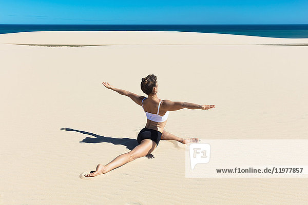 Rückansicht einer Frau am Strand beim Spagat,  Arme in Yogastellung geöffnet