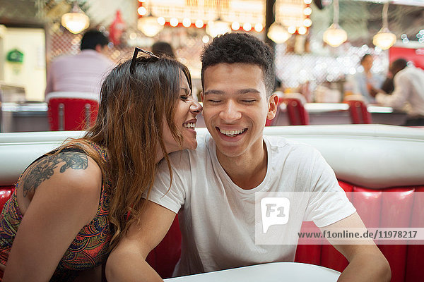 Junges Paar sitzt im Diner,  junge Frau flüstert dem Mann ins Ohr und lacht