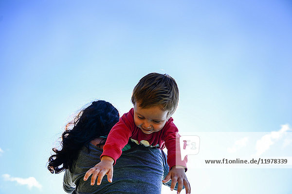 Low angle view of male toddler over mother's shoulder against blue sky