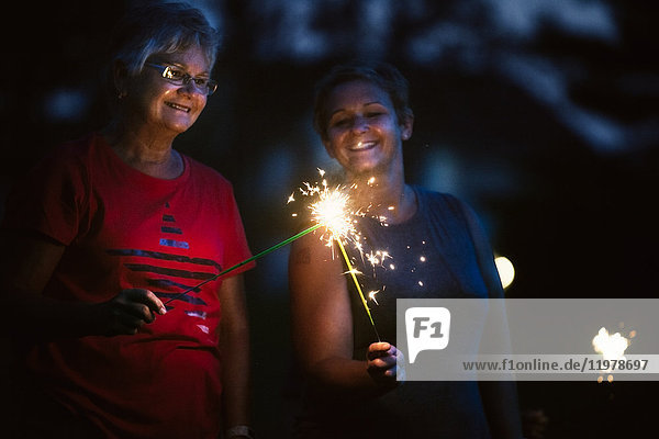 Senior and mature woman igniting sparklers together at night on independence day  USA