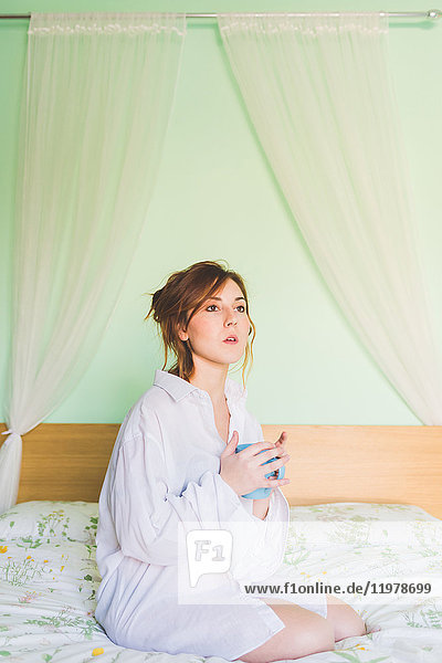 Young woman kneeling on bed holding coffee cup and staring