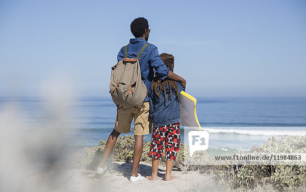Vater und Tochter mit Boogie-Board mit Blick auf den Ozean am sonnigen Sommerstrand