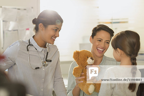 Lächelnde Kinderärztin und Mutter zeigen einem Mädchen im Untersuchungsraum einen Teddybären