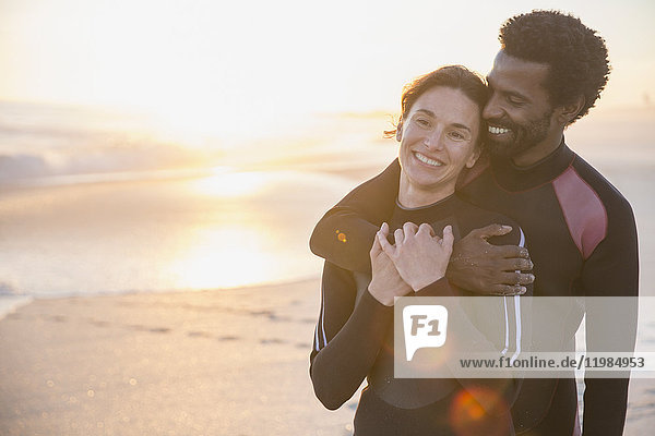 Lächelndes  liebevolles multiethnisches Paar in Neoprenanzügen am sonnigen Sommer-Sonnenuntergangsstrand.