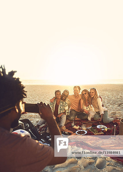 Young man with camera phone photographing friends enjoying picnic on sunny summer beach