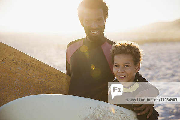 Portrait lächelnder,  selbstbewusster Vater und Sohn in Neoprenanzügen mit Surfbrettern am sonnigen Sommerstrand.