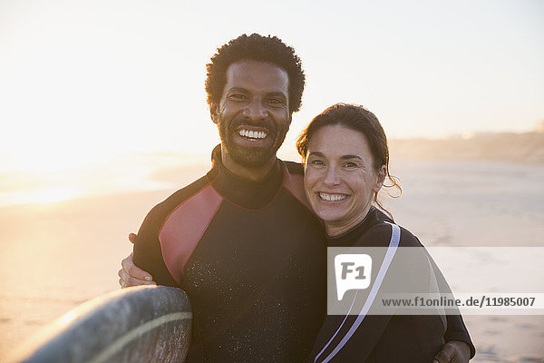 Portrait lächelndes  selbstbewusstes multiethnisches Paar mit Surfbrett am sonnigen Sommersonnenuntergangstrand.