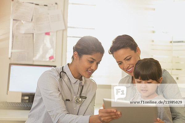 Eine Kinderärztin zeigt einer Patientin und ihrer Mutter im Untersuchungsraum ein digitales Tablet
