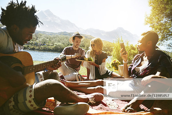 Junge Freunde hängen herum  spielen Gitarre und genießen ein Picknick am sonnigen Sommerufer des Flusses.