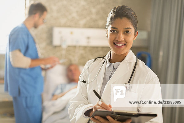 Porträt einer lächelnden Ärztin bei der Visite  die sich Notizen auf einem Klemmbrett in einem Krankenhauszimmer macht