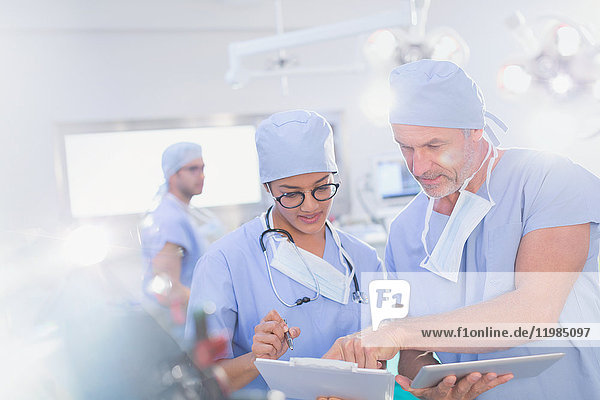 Chirurgen mit digitalem Tablet besprechen Papierkram auf einem Klemmbrett im Operationssaal