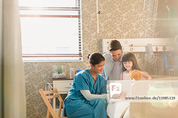 Female nurse showing digital tablet to girl patient and mother in hospital room